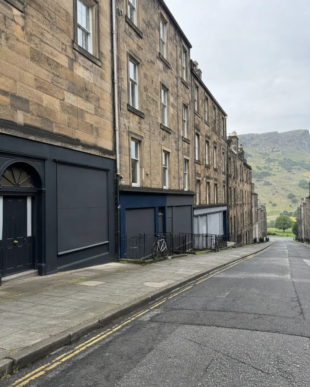 Stone tenements above blank painted shopfronts on a sloping Southside street, Salisbury Crags beyond