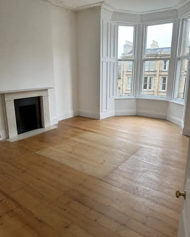 An empty bay-windowed living room with a marble fireplace and a pale patch where a rug sat